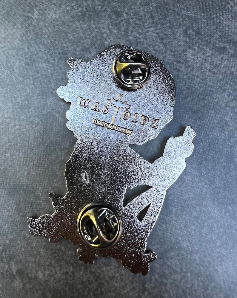 May include: A silver-colored enamel pin with a cartoon figure giving the middle finger. The pin has two pin backs and the text "WAS TOIDZ" and "WARTOIDZ.COM" is visible.