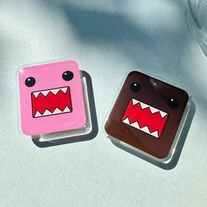 May include: Two square Domo-kun character stickers. One is pink, the other is brown. Both have black eyes and a wide, toothy mouth with red and white details. The stickers are on a light blue surface.