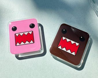 以下が含まれることがあります： 2つの正方形のドモ君キャラクターのステッカー。1つはピンク、もう1つは茶色です。どちらも黒い目と、赤と白のディテールが施された、幅広で歯のある口があります。ステッカーは水色の表面にあります。