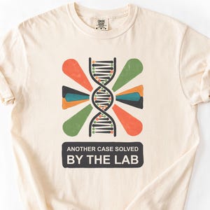 Puede incluir: Camiseta de color marfil con un gráfico colorido de una hélice de ADN con detalles en naranja, verde y azul. El texto "ANOTHER CASE SOLVED BY THE LAB" está impreso debajo en un rectángulo negro.