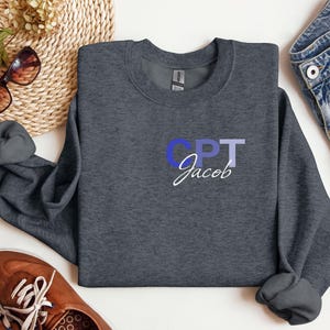 May include: A dark heather gray sweatshirt with the text "CPT Jacob" in white and blue lettering.