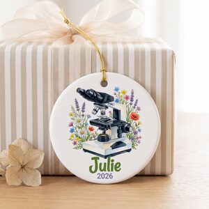 Op de afbeelding: Een rond wit keramieken ornament met een zwart-witte microscoopillustratie omringd door kleurrijke wilde bloemen. De naam Julie staat in groene tekst boven het jaartal 2026 in paarse tekst. Aan de bovenkant zit een gouden koordje.