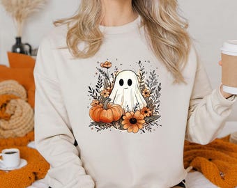 May include: A cream-colored sweatshirt featuring a Halloween-themed graphic. The design includes a friendly ghost, pumpkins, and flowers in shades of orange, brown, and green. The ghost has a simple, smiling face. The sweatshirt is a casual, long-sleeved garment.