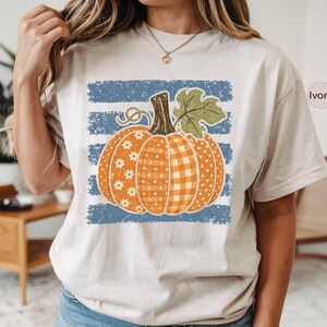 May include: An ivory short-sleeve t-shirt featuring a graphic of a pumpkin with orange floral, plaid, and polka dot patterns. The pumpkin is set against a distressed blue horizontal striped background. The word Ivory is printed in a small circle.