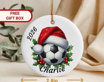 Op de afbeelding: Een rond wit keramieken ornament met een diameter van 7,1 cm, voorzien van een voetbal met een rode kerstmuts, omringd door groene hulstbladeren en rode bessen. De tekst 2026 en de naam Charlie staan op het ornament gedrukt.