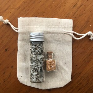 May include: Two small glass jars with cork tops, one filled with white sage and the other with brown crystals, are sitting inside a white drawstring bag.