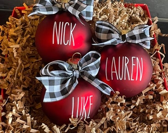 May include: Three red glass ornaments with black and white checkered ribbon bows. The ornaments are personalized with the names "Nick", "Lauren", and "Luke" in white lettering.