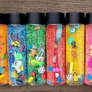 May include: Six clear sensory bottles filled with colored rice and small toys. The bottles have black lids and contain orange, blue, green, red, yellow, and pink rice. Toys include a skateboard, dice, and animal figurines.