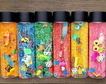 Può includere: Sei bottiglie sensoriali trasparenti riempite con riso colorato e piccoli giocattoli. Le bottiglie hanno coperchi neri e contengono riso arancione, blu, verde, rosso, giallo e rosa. I giocattoli includono uno skateboard, dadi e figurine di animali.