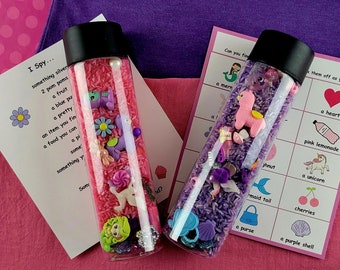 Può includere: Due bottiglie sensoriali trasparenti riempite con riso colorato e piccoli giocattoli. Una bottiglia ha riso rosa e l'altra viola. Sono visibili schede con richieste "I Spy" e "Can you find".