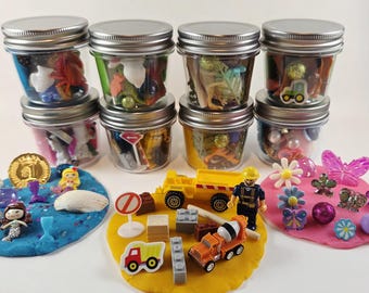 Può includere: Otto barattoli di vetro trasparente con coperchi argentati riempiti di piccoli giocattoli e ninnoli di plastica. Sotto i barattoli ci sono tre mucchi di pasta modellabile blu, gialla e rosa decorati con miniature, veicoli, fiori e gemme.