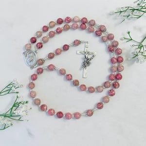 May include: A pink and white stone rosary with a silver crucifix and a silver medal of the Virgin Mary. The rosary is laid out on a white surface.