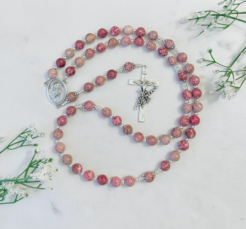 May include: A pink and white stone rosary with a silver crucifix and a silver medal of the Virgin Mary. The rosary is laid out on a white surface.
