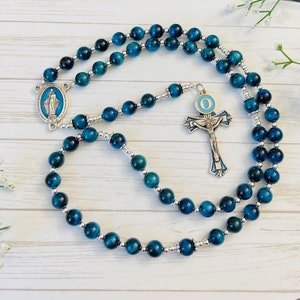 May include: Blue tiger's eye rosary beads with a silver crucifix and a silver charm of the Virgin Mary. The crucifix has a blue and gold enamel design with the letter "O" on the center circle.