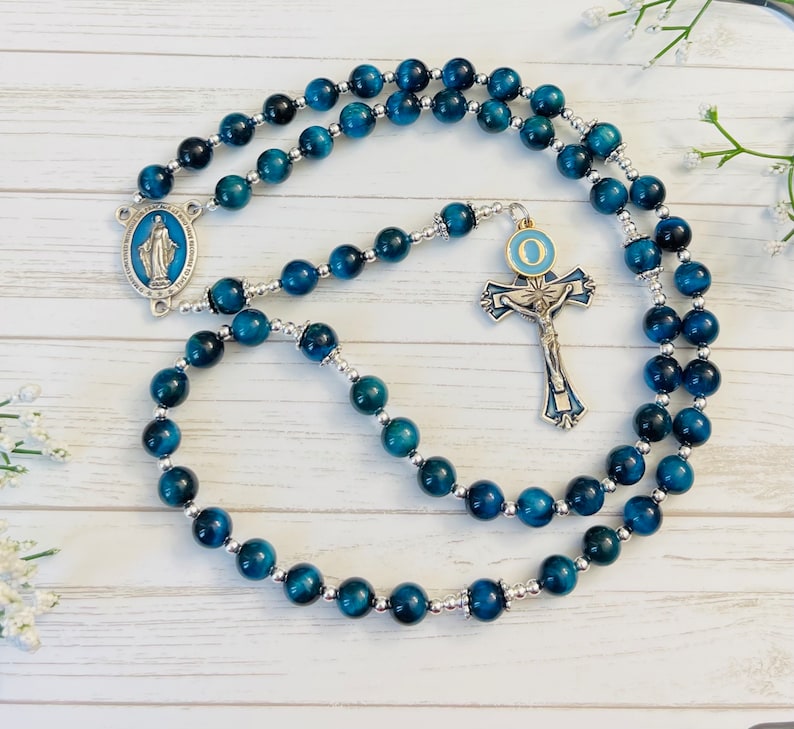 May include: Blue tiger's eye rosary beads with a silver crucifix and a silver charm of the Virgin Mary. The crucifix has a blue and gold enamel design with the letter "O" on the center circle.