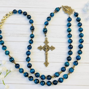 May include: A blue tiger's eye rosary with gold beads and a brass crucifix. The rosary has a personalized name tag that reads "REGINA".