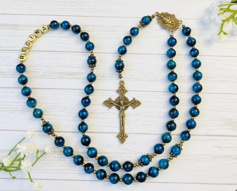 May include: A blue tiger's eye rosary with gold beads and a brass crucifix. The rosary has a personalized name tag that reads "REGINA".