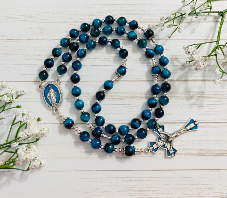 May include: Blue tiger's eye bead rosary with silver accents. The rosary features a silver pendant with a blue image of the Virgin Mary and a silver crucifix with a blue image of Jesus.