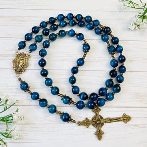 May include: A blue tiger's eye rosary with a brass crucifix and a brass medal of the Virgin Mary. The rosary is laid out on a white wooden surface.