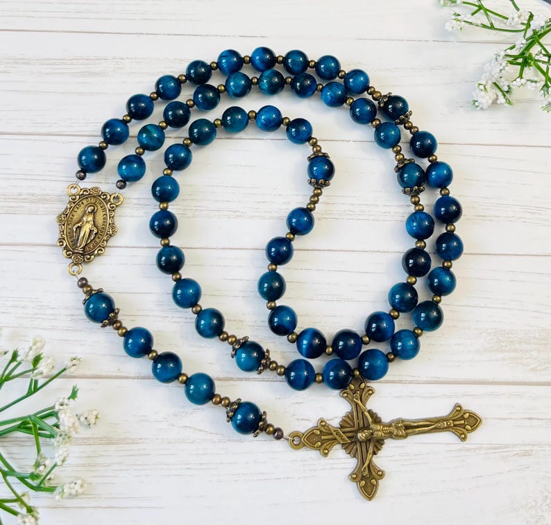 May include: A blue tiger's eye rosary with a brass crucifix and a brass medal of the Virgin Mary. The rosary is laid out on a white wooden surface.