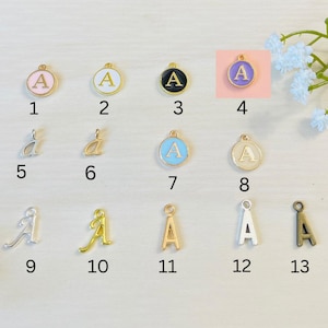 May include: Thirteen different charms featuring the letter "A" in various styles and colors. The charms are arranged in three rows of four, with one charm in the bottom right corner. The charms are made of metal and have a gold finish. Some charms are round with a white, pink, blue, or cream enamel background. Other charms are a cursive letter "A" in silver, gold, or bronze.