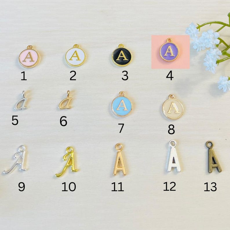 May include: Thirteen different charms featuring the letter "A" in various styles and colors. The charms are arranged in three rows of four, with one charm in the bottom right corner. The charms are made of metal and have a gold finish. Some charms are round with a white, pink, blue, or cream enamel background. Other charms are a cursive letter "A" in silver, gold, or bronze.