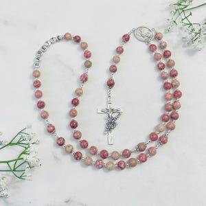 May include: A pink and beige stone rosary with a silver cross and a silver charm featuring the Virgin Mary. The rosary beads are personalized with the name "BABETTE".