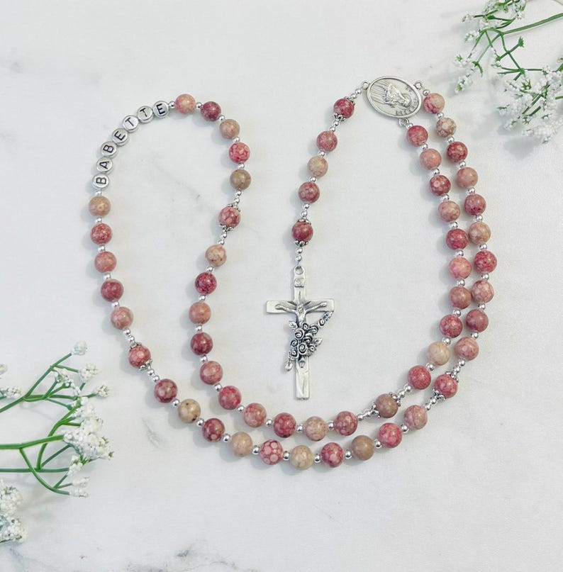May include: A pink and beige stone rosary with a silver cross and a silver charm featuring the Virgin Mary. The rosary beads are personalized with the name "BABETTE".
