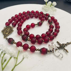May include: A red beaded rosary with a detailed crucifix and a pendant featuring a religious figure. The beads are a deep red colour, and the metal accents have a vintage bronze finish. The rosary is displayed on a white surface.