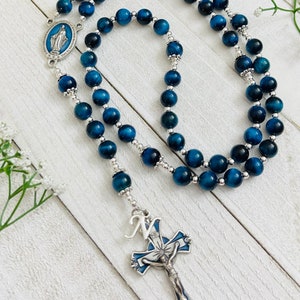 May include: Blue tiger's eye bead rosary with a silver crucifix and a silver charm featuring the Virgin Mary. The crucifix has a blue and white enamel design and the letter "N" on the top.