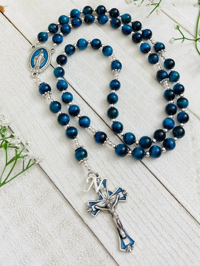 May include: Blue tiger's eye bead rosary with a silver crucifix and a silver charm featuring the Virgin Mary. The crucifix has a blue and white enamel design and the letter "N" on the top.