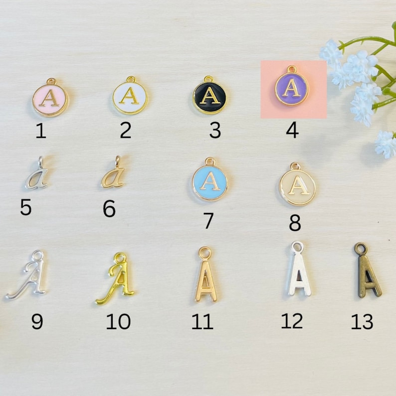 May include: Thirteen different charms featuring the letter 'A' in various styles and colors. The charms are arranged in three rows of four, with one charm in the bottom right corner. The charms are made of metal and have a gold finish. Some charms are round, while others are more stylized. The charms are numbered 1 through 13.