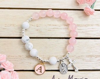 May include: A pink and white beaded bracelet with a silver charm that says "Ave Maria" and a gold charm with the letter "A".