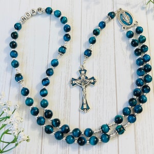 May include: Blue and silver rosary beads with a silver cross and a silver charm with a blue image of the Virgin Mary. The beads are made of tiger's eye stone. The rosary is personalized with the name "Noah".
