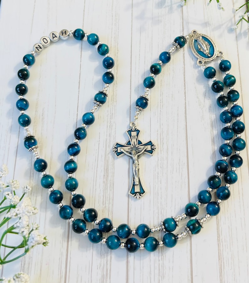 May include: Blue and silver rosary beads with a silver cross and a silver charm with a blue image of the Virgin Mary. The beads are made of tiger's eye stone. The rosary is personalized with the name "Noah".