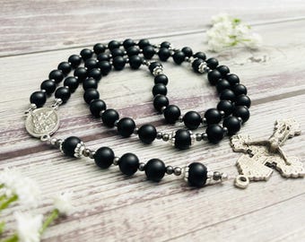 Puede incluir: Cuentas de rosario de ónix negro con un crucifijo de plata y un dije de plata con una imagen religiosa. Las cuentas están enhebradas en una cadena de plata.