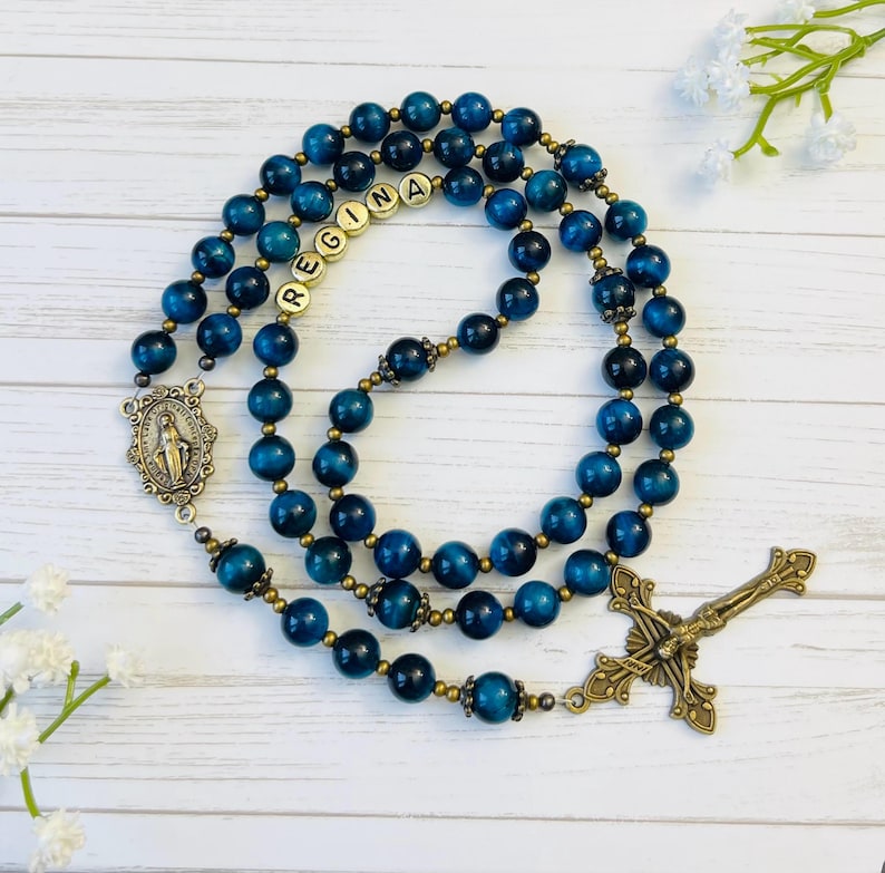 May include: Blue and gold rosary beads with a brass crucifix and a charm of the Virgin Mary. The beads are made of tiger's eye stone and the name "REGINA" is spelled out with gold letter beads.