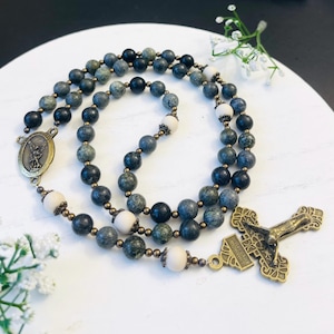May include: A rosary with dark gray and white beads. The beads are strung together with a bronze-colored chain. The rosary has a bronze-colored crucifix and a medal with an image of a saint.
