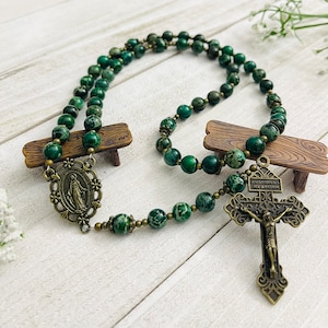 May include: Green and gold rosary beads with a green and gold crucifix and a green and gold medal of the Virgin Mary. The rosary is laid out on a white wooden surface.