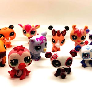 May include: A collection of colorful, small animal figurines with large eyes. The figurines include a variety of animals such as pandas, deer, and bears, in shades of orange, pink, white, and purple. The toys are arranged on a white surface.