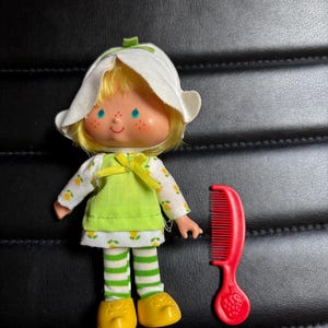 May include: A vintage doll with blonde hair, blue eyes, and freckles, wearing a white bonnet, a green dress, and striped green and white leggings. A red comb with a strawberry design is next to the doll. The doll's yellow shoes complete the outfit.