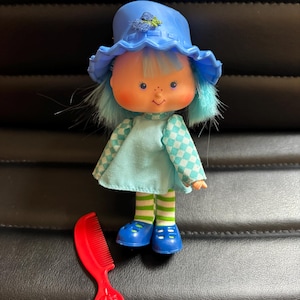 May include: A vintage doll with blue hair, a matching hat, and a light blue dress. The doll has a red comb with a strawberry design. The doll is wearing blue shoes and green and white striped socks. The doll is a collectible item.