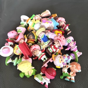 May include: A collection of small, colorful figurines featuring characters in various poses. The figurines have a cartoonish style, with bright colors and detailed outfits. Some figurines are holding accessories. The collection is on a dark surface.