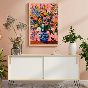 May include: A white wooden cabinet with black hairpin legs, a white vase with green leaves, a silver vase with dried flowers, and a framed painting of a blue and white vase filled with colorful flowers on a pink background.