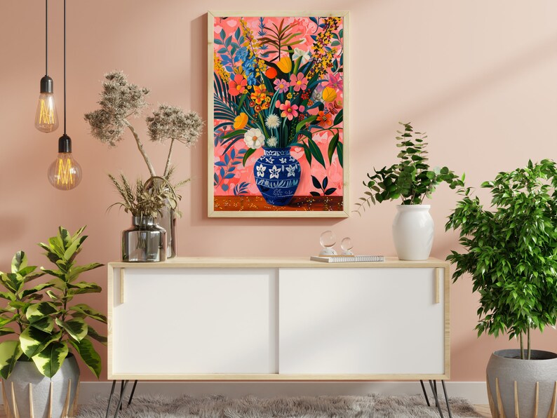 May include: A white wooden cabinet with black hairpin legs, a white vase with green leaves, a silver vase with dried flowers, and a framed painting of a blue and white vase filled with colorful flowers on a pink background.