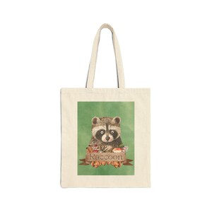 May include: A beige tote bag with a green background and a cartoon raccoon with a brown and gray fur pattern. The raccoon is holding a small basket of fruit and nuts. The text "Raccoon" is written in brown letters on a wooden sign.