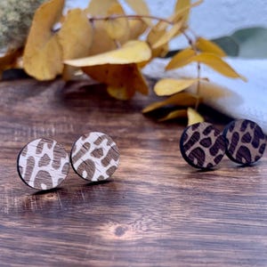 Könnte beinhalten: Zwei Paar Holz-Ohrstecker mit Leopardenmuster. Ein Paar hat einen hellen Holzgrund mit braunen Flecken, das andere einen dunklen Holzgrund mit braunen Flecken. Die Ohrringe sind rund und liegen auf einer Holzoberfläche.