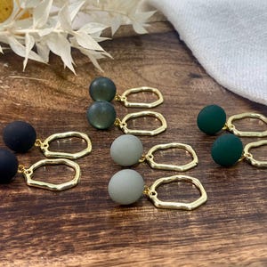 Könnte beinhalten: Ohrringe mit matten Kugeln in Schwarz, Grün und Grau, kombiniert mit goldfarbenen, unregelmäßig geformten Reifen. Die Ohrringe sind auf einer dunklen Holzoberfläche angeordnet, mit einem weißen Tuch und getrockneten floralen Akzenten.