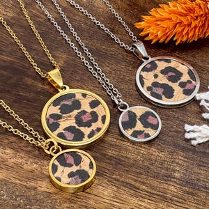 Könnte beinhalten: Vier Halsketten mit runden Anhängern mit Leopardenmuster. Zwei Halsketten haben goldene Ketten und Lünetten, während die anderen beiden silberne Ketten und Lünetten haben. Die Anhänger sind aus Kork mit schwarzen und rosa Flecken.