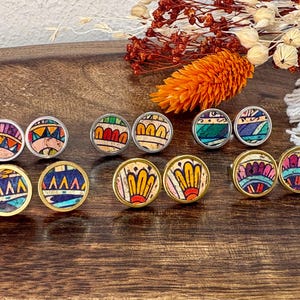 Könnte beinhalten: Eine Sammlung runder Ohrstecker mit farbenfrohen, gemusterten Designs. Die Ohrringe weisen eine Vielzahl von Farben auf, darunter Rot, Blau, Gelb und Grün, die in silber- und goldfarbenen Metalleinfassungen gefasst sind. Die Ohrringe werden auf einer Holzoberfläche präsentiert.